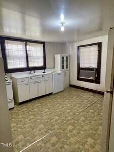 A small kitchen with patterned linoleum flooring, white cabinets, a sink, a stove, a compact refrigerator, and a glass-front cabinet. Two windows with blinds and a window air conditioner unit are visible.