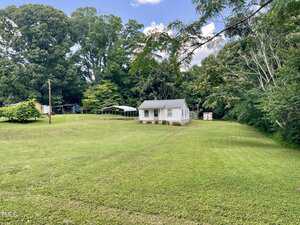 A small white house sits in the middle of a large grassy yard with scattered trees and a detached carport. The sky is partly cloudy, and there are woods in the background.