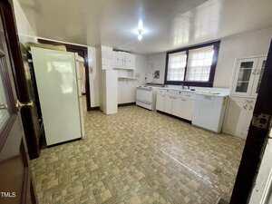 A spacious kitchen with vinyl tile flooring, white cabinets, a refrigerator, a stove, a sink under a window with blinds, and a small wall cabinet. The room is lit by a single ceiling light.
