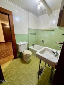 A small bathroom with green tile floors and walls, a white toilet, a white sink with exposed pipes, and a bathtub. A towel rack is above the tub, and a single light bulb hangs from the ceiling.