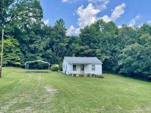 Nestled at 229 Maple Drive in Oxford, this small white house with a front porch overlooks a large, well-kept lawn. A metal carport frame sits to the left, while dense trees and greenery create a scenic backdrop beneath a partly cloudy blue sky.