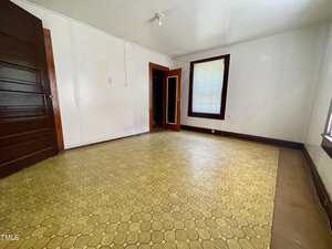 Empty room with yellow hexagonal patterned linoleum flooring, white walls, a dark wooden door, a closet with an open door, and two windows with dark wood trim and one partially closed blind.