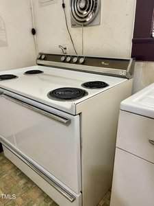 A white electric stove with four coil burners and a control panel with knobs is positioned next to a washing machine in a kitchen with patterned linoleum flooring. An exhaust fan is installed on the wall above.