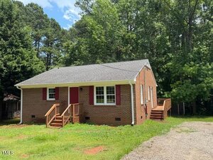 A small, single-story brick house at 818 Wakefield Avenue, Henderson, features red shutters and doors, wooden steps to the front and side entrances, and is surrounded by trees and a grassy yard under a partly cloudy sky.