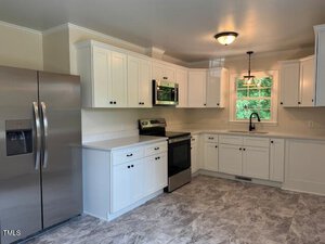 A modern kitchen with white cabinets, stainless steel appliances including a refrigerator and stove, a microwave, a single window above the sink, and gray tiled flooring.
