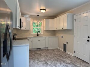 A kitchen with white cabinets, stainless steel appliances, a single window above the sink, a ceiling light fixture, and light gray patterned flooring. There is a white door on the right side of the image.