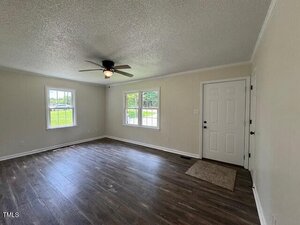 A vacant living room with light-colored walls, dark wood flooring, two windows, a white front door, a beige doormat, and a ceiling fan with a light fixture. Natural light is coming through the windows.
