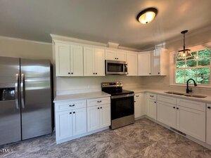 A kitchen with white cabinets, stainless steel appliances including a refrigerator, oven, and microwave, a single-basin sink under a window, overhead lighting, and a tiled floor.