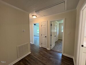 A hallway with wood flooring, beige walls, white trim, and several white doors leading to different rooms; an attic access panel is visible in the ceiling. One room has carpet, another has light-colored floors.