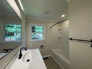 A bathroom with a white sink, black faucet, large mirror, white bathtub with shower, black towel bar, and two windows showing trees outside. The walls are light-colored and the floor has a tile pattern.