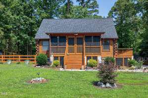 A log cabin with a stone foundation, screened-in porch, and wooden stairs sits in a grassy yard at 52 Buoy Drive, Henderson, surrounded by shrubs and trees on a sunny day.