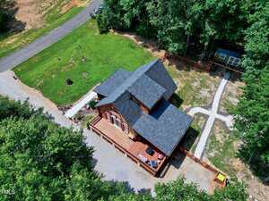 Aerial view of a wooden house with a dark roof, spacious deck, and surrounding gravel and grass areas. A driveway leads to the main road, and there is a small outbuilding near the trees at the back.