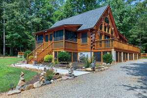 A log cabin with a screened porch, large wooden deck, and garden beds in front. The house is surrounded by trees and a gravel driveway with decorative rock arrangements lining the walkway.