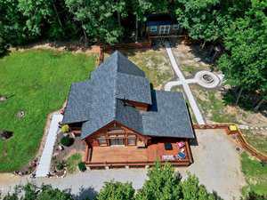 Aerial view of a wooden cabin with a large deck, surrounded by grass, trees, and a gravel driveway. A separate small building and a fire pit area are visible in the fenced backyard.