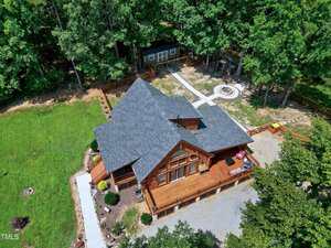 Aerial view of a wooden house with a large deck, surrounded by trees and fenced yard. A gravel driveway leads to the entrance, and a fire pit area with seating is visible in the backyard.