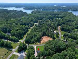 Aerial view of a wooded residential area with winding roads, scattered houses, and a cleared lot. A red location marker highlights a specific property. A large lake is visible in the background.
