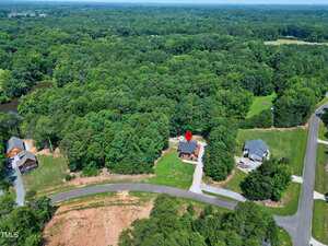 Aerial view of a house marked with a red location pin, surrounded by trees and greenery. Two neighboring houses are visible, with a main road curving through the bottom of the image.