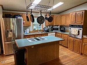 A kitchen with wooden cabinets, a stainless steel refrigerator, an island with a stovetop, hanging pans, a double oven, a dishwasher, and various small appliances on the counters. A window is above the sink.