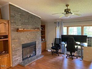 A living room with a stone fireplace, wooden floors, a ceiling fan, built-in wooden shelves, and a home office setup with two computer monitors by a window.