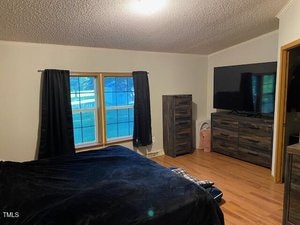 A bedroom with light-colored walls and wood flooring, featuring a bed with dark bedding, a large window with black curtains, a dresser, a tall chest of drawers, a large wall-mounted TV, and a laundry basket.