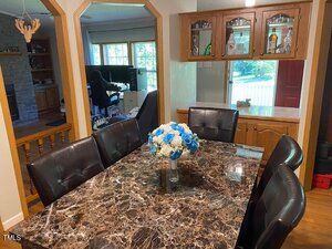 A dining room with a dark marble table, six black cushioned chairs, and a vase of blue and white flowers. In the background are a home office, a living room, and a wooden china cabinet.