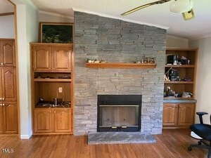 A room with a stone fireplace centered between two built-in wooden cabinets and shelves. Items are placed on the shelves. The floor is hardwood, and a ceiling fan is partially visible at the top right.