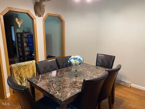A dining room with a dark marble table, six black cushioned chairs, and a vase of white and blue flowers. The room has hardwood floors, light walls, and two arched doorways with wood trim. A mounted deer head is on the wall.