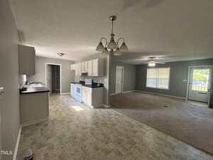 Open-plan kitchen and living room with light gray walls, white cabinets, and blue appliances. The kitchen has tile flooring, while the living area is carpeted. Large windows let in natural light.