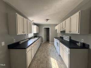 A kitchen with white cabinets, dark countertops, light-colored tile flooring, a window over the sink, and appliances including a stove and range hood. The walls are painted gray and there is a door at the end of the room.