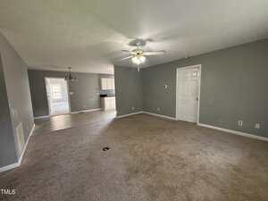 A spacious living room with beige carpet, light gray walls, and a ceiling fan. The room opens to a tiled dining area and a kitchen with white cabinets. Multiple doors are visible along the walls.