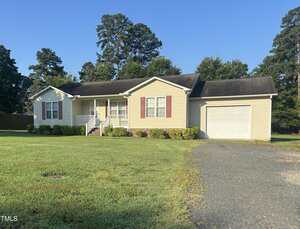 A charming single-story beige house at 607 24th Street in Butner features red shutters, a white garage door, and sits on a grassy lawn with a gravel driveway. Bushes line the front, with tall trees providing a scenic backdrop.