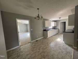 A kitchen with light-colored cabinets, black countertops, and vinyl flooring. A chandelier hangs from the ceiling, and an adjacent carpeted room is visible through an open doorway. Sunlight enters through a window.