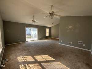 A large empty living room with gray walls, two ceiling fans, beige carpet, and several wall patches. Sunlight comes through windows and a sliding glass door that leads to a deck outside.