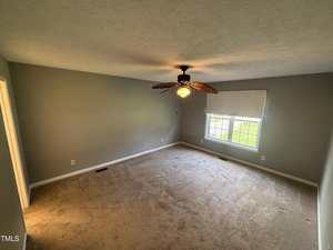 Empty carpeted room with gray walls, a ceiling fan with leaf-shaped blades, a large window with a white blind, and natural light coming in. Electrical outlets and a vent are visible along the walls.