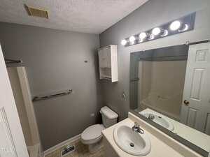 Small bathroom with gray walls, a white toilet, white sink, white medicine cabinet, and mirror with a row of five lights above. Tiled floor, shower-tub combo, towel bar, and air vent visible. Door partly open.