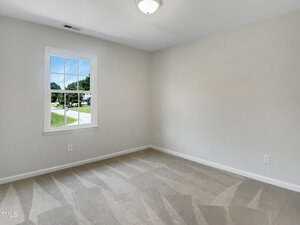 An empty room with beige carpet, light gray walls, a single window showing a street and green trees outside, and a ceiling light fixture. The space appears clean and well-lit.
