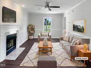 A living room with white walls, dark flooring, and a ceiling fan. It features a sofa, armchair, coffee table, decorative plants, and a fireplace with a mounted TV above. The image is labeled "Virtually Staged.