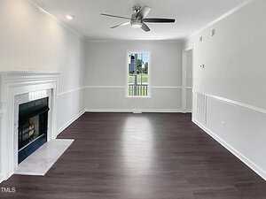 A bright, empty living room with white walls, dark wood flooring, a fireplace on the left, a ceiling fan with lights, and a window in the center at the far end letting in natural light.