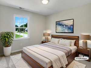 A neatly made bed with white and beige bedding sits against a light-colored wall in a bedroom. Two nightstands with lamps, a potted plant, a window, and a framed picture are visible. The image is labeled "Virtually Staged.