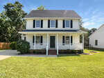A two-story white house with black shutters and a covered front porch at 1599 Rogers Pointe Lane, Creedmoor, sits on a well-manicured lawn with shrubs and a paved driveway. Trees are visible in the background under a blue sky.