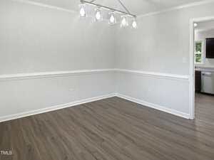 An empty room with light gray walls, white trim, wood-look flooring, and a modern ceiling light fixture. An open doorway leads to a kitchen with visible stainless steel appliances.