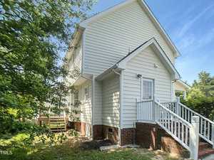 Two-story white house with horizontal siding, red brick foundation, and white steps leading to a back door. There are mature trees and green grass surrounding the property. The sky is clear and blue.