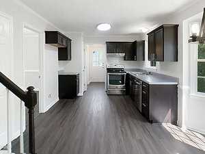 A modern kitchen with dark wood cabinets, stainless steel appliances, dark countertops, and wood-look flooring. Natural light enters from a window by the sink and a glass-paned door at the back.