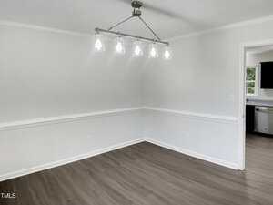 A bright, empty room with white walls, crown molding, wood-style flooring, and a modern five-light ceiling fixture. An open doorway leads to a kitchen with stainless steel appliances.