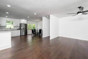 A modern, open-concept kitchen and dining area with white cabinets, stainless steel appliances, dark wood flooring, and large windows; an adjacent empty living space has a ceiling fan.