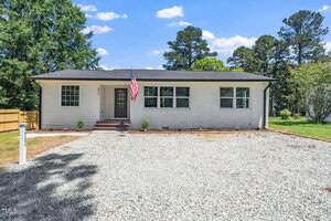 Charming single-story white brick house at 311 Troy Street, Creedmoor, with a black roof, American flag by the door, several windows, small shrubs in front, and a gravel driveway. Surrounded by trees and a wooden fence on the left.