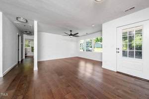 A spacious, empty living room with white walls, wood flooring, large windows, a ceiling fan, recessed lighting, and a glass door leading outside. A hallway is visible to the left.