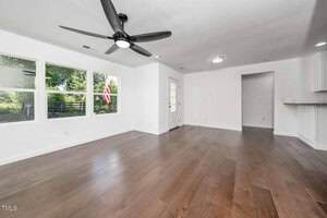 A bright, empty living room with white walls, dark wood flooring, three large windows, a ceiling fan, overhead lights, and a white door leading outside. Part of a kitchen counter is visible on the right.