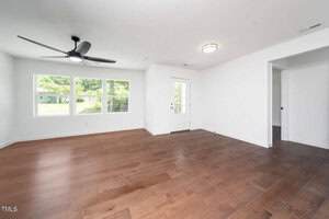 A bright, empty room with white walls, three large windows, a ceiling fan, wooden floors, and a glass door leading outside. An open doorway is visible on the right side of the room.