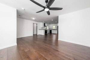 A spacious room with dark wood flooring, white walls, a black ceiling fan, and recessed lighting. The open layout leads to a modern kitchen with white cabinets and stainless steel appliances.